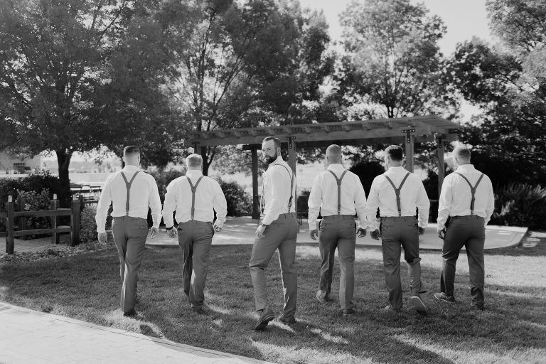 Six men in suspenders walk away outdoors, near trees and a pergola. Black and white photo.