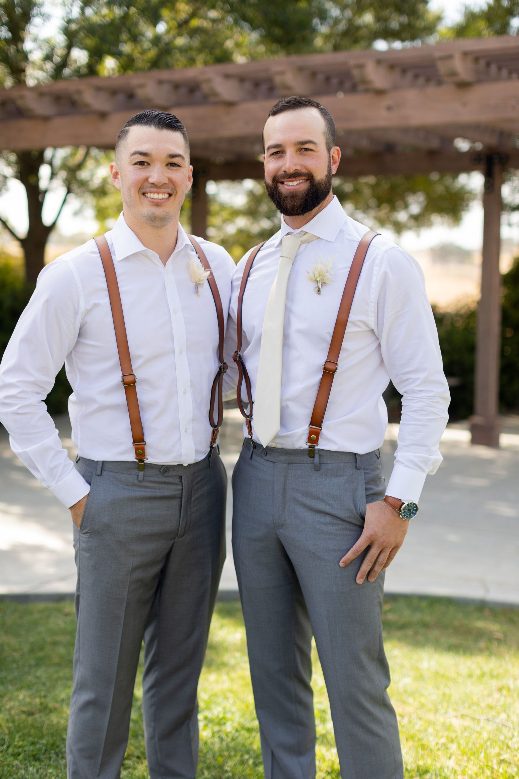 Two men in dress shirts and suspenders smile outdoors.