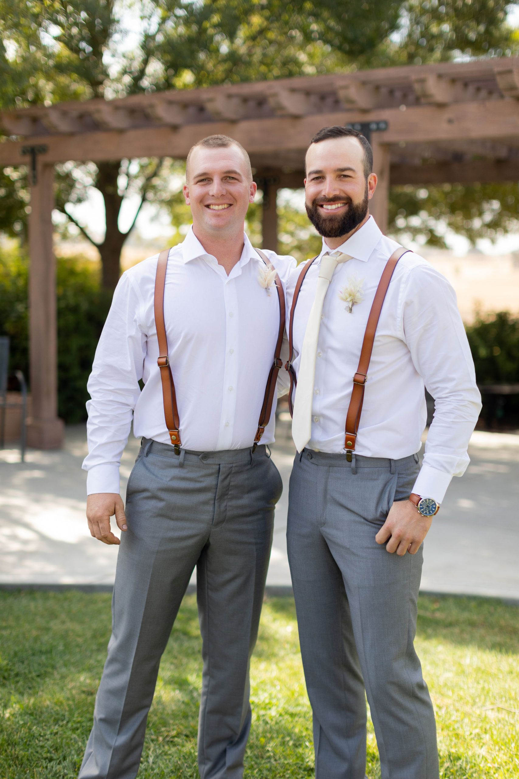 Two men in white shirts, grey pants, and brown suspenders stand outside smiling.
