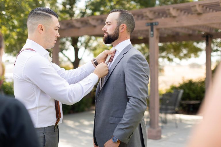 Man adjusts other man's tie. Both wear suits, outdoors by a pergola.