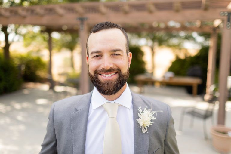 Man in gray suit and tie smiles, with a full beard, outdoors near a wooden arbor.