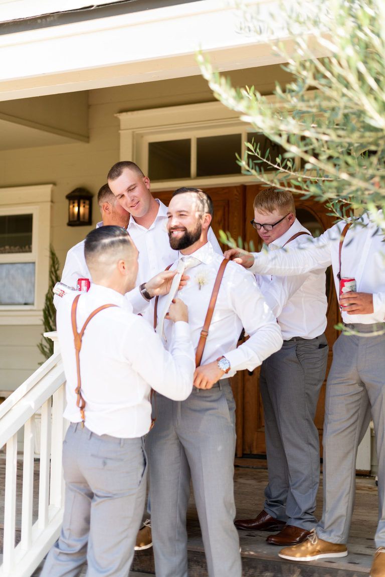 Groom with groomsmen adjusting tie and suspenders on a porch. Men wear white shirts, grey pants, and brown suspenders.