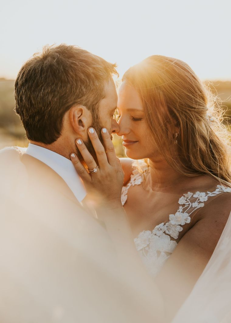 Bride and groom embrace, smiling. Bride in white dress, groom in blue suit, standing outdoors with vineyard background.
