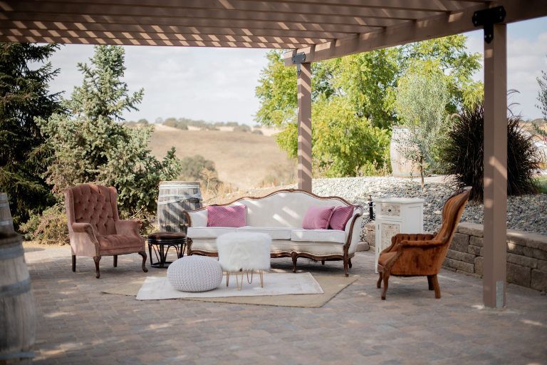 Outdoor lounge area: white couch with pink pillows, two armchairs, small tables, under a wooden pergola, landscape background.