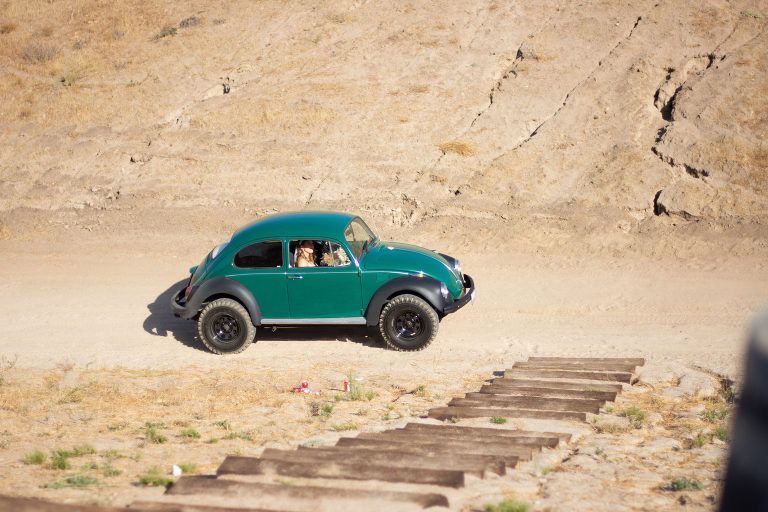 Green VW Beetle off-roading on a dirt path with passengers, set against a brown hillside.