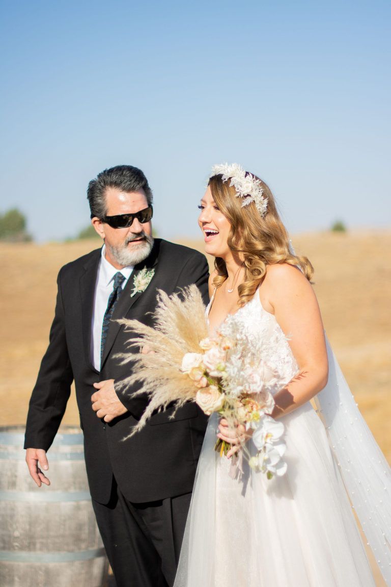 Bride laughing, walking with her father down an outdoor aisle. She wears a white dress and flower crown, he wears a suit.