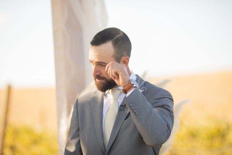 Groom wiping a tear during outdoor wedding ceremony; wearing a gray suit and cream tie.