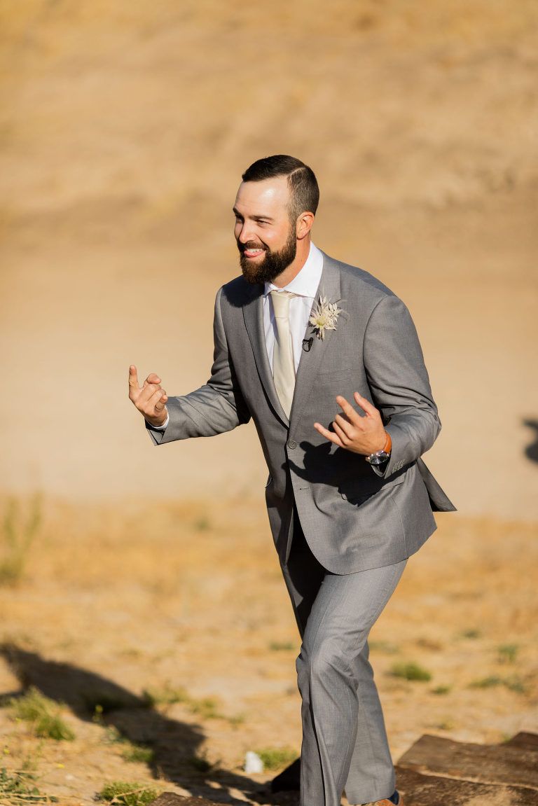 Man in gray suit with hands up, smiling, outdoors.