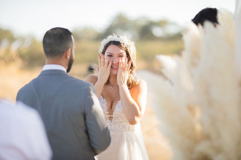 Bride wiping tears, facing groom, at outdoor wedding ceremony.