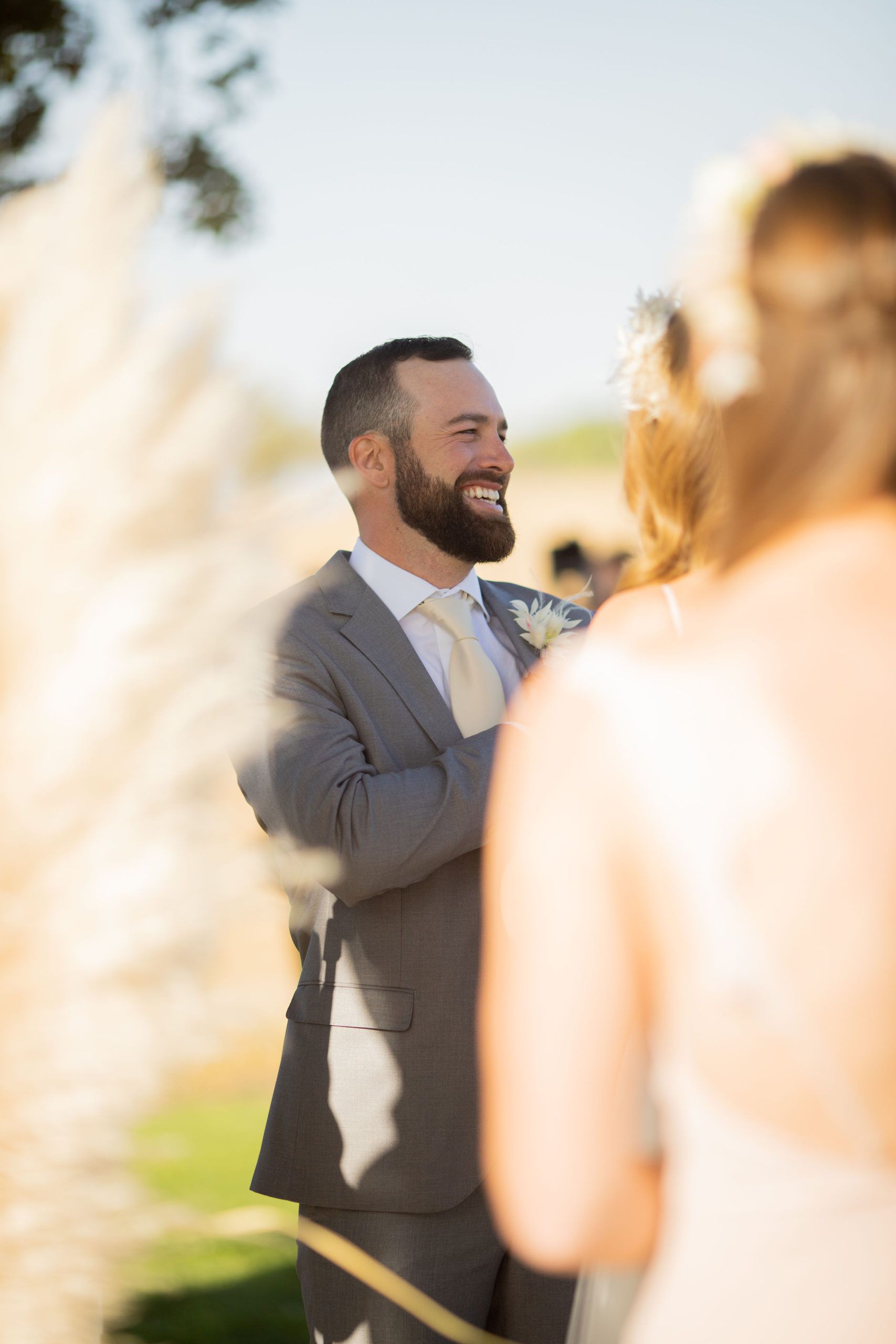 Groom in gray suit smiles during outdoor wedding ceremony. Bride's back is to camera. Pampas grass in foreground.