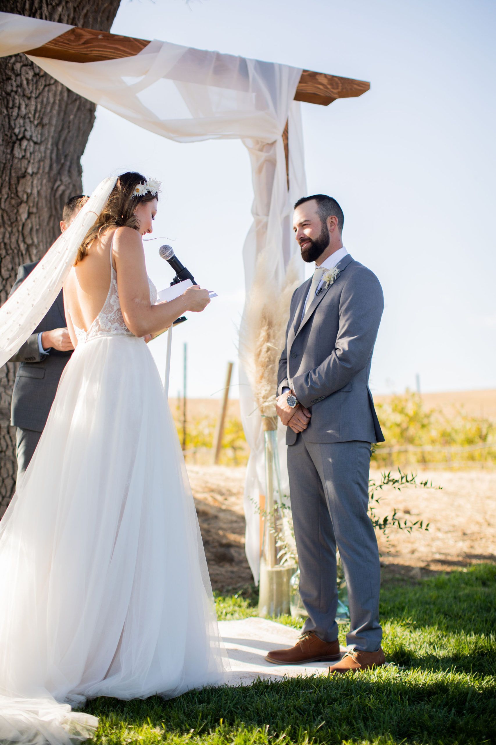 Bride reading vows to groom during outdoor wedding ceremony. Wooden arch decorated with white fabric.