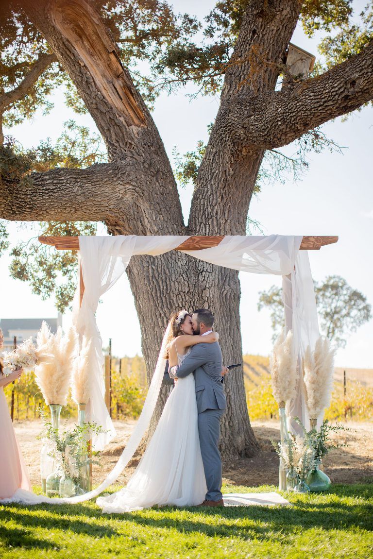Bride and groom embrace under a wooden arch decorated with fabric and pampas grass during an outdoor wedding ceremony.