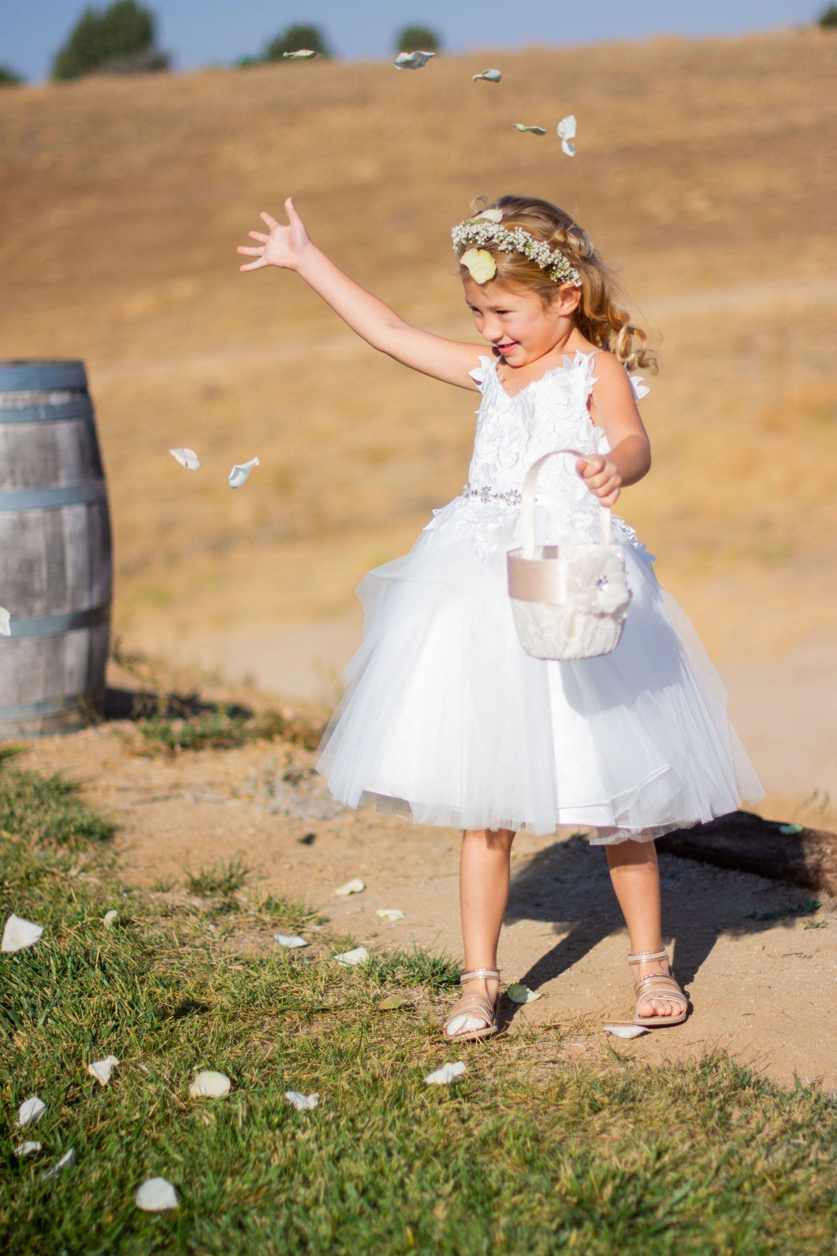 Girl in white dress throws flower petals, smiling outdoors.