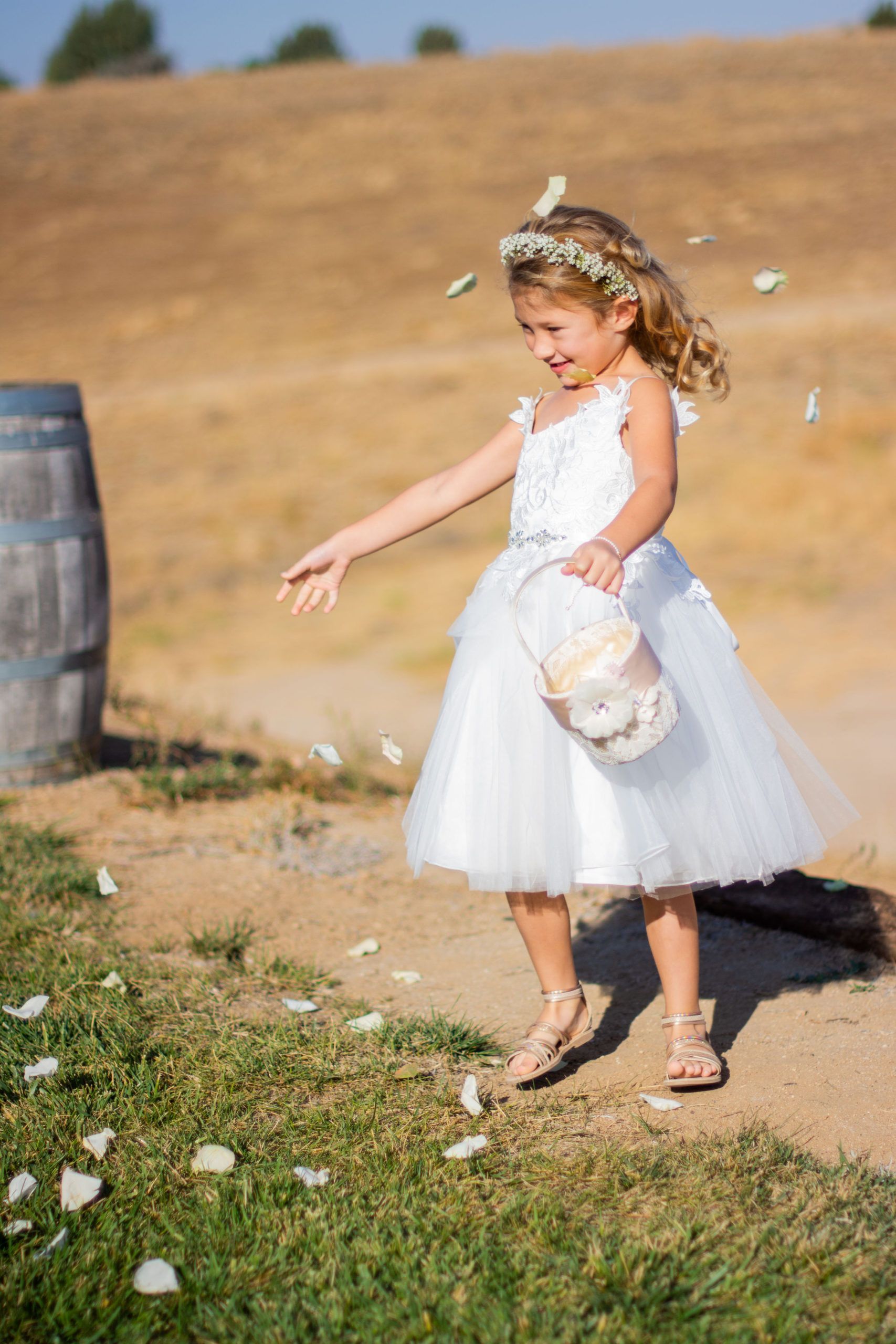 Young girl in white dress, scattering flower petals at an outdoor wedding.