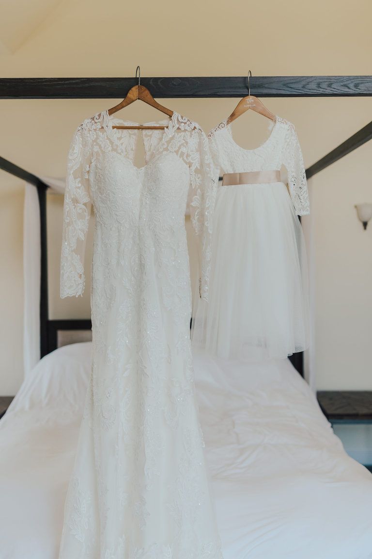 Wedding dress and flower girl dress hanging on a bed.