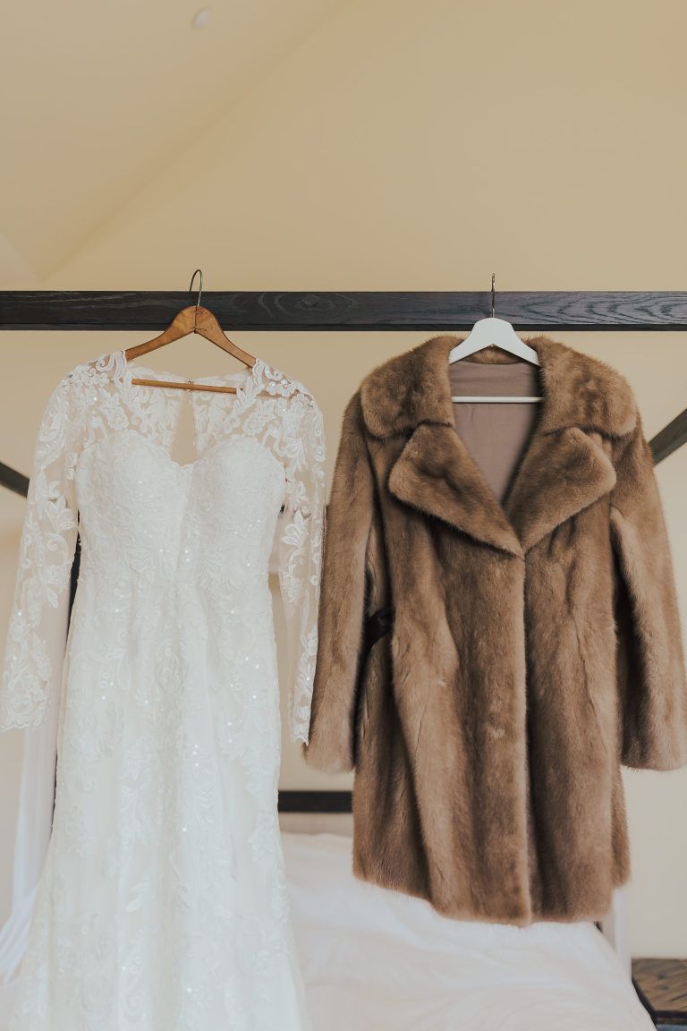 Wedding dress with long lace sleeves and a brown fur coat hanging from a wooden beam.