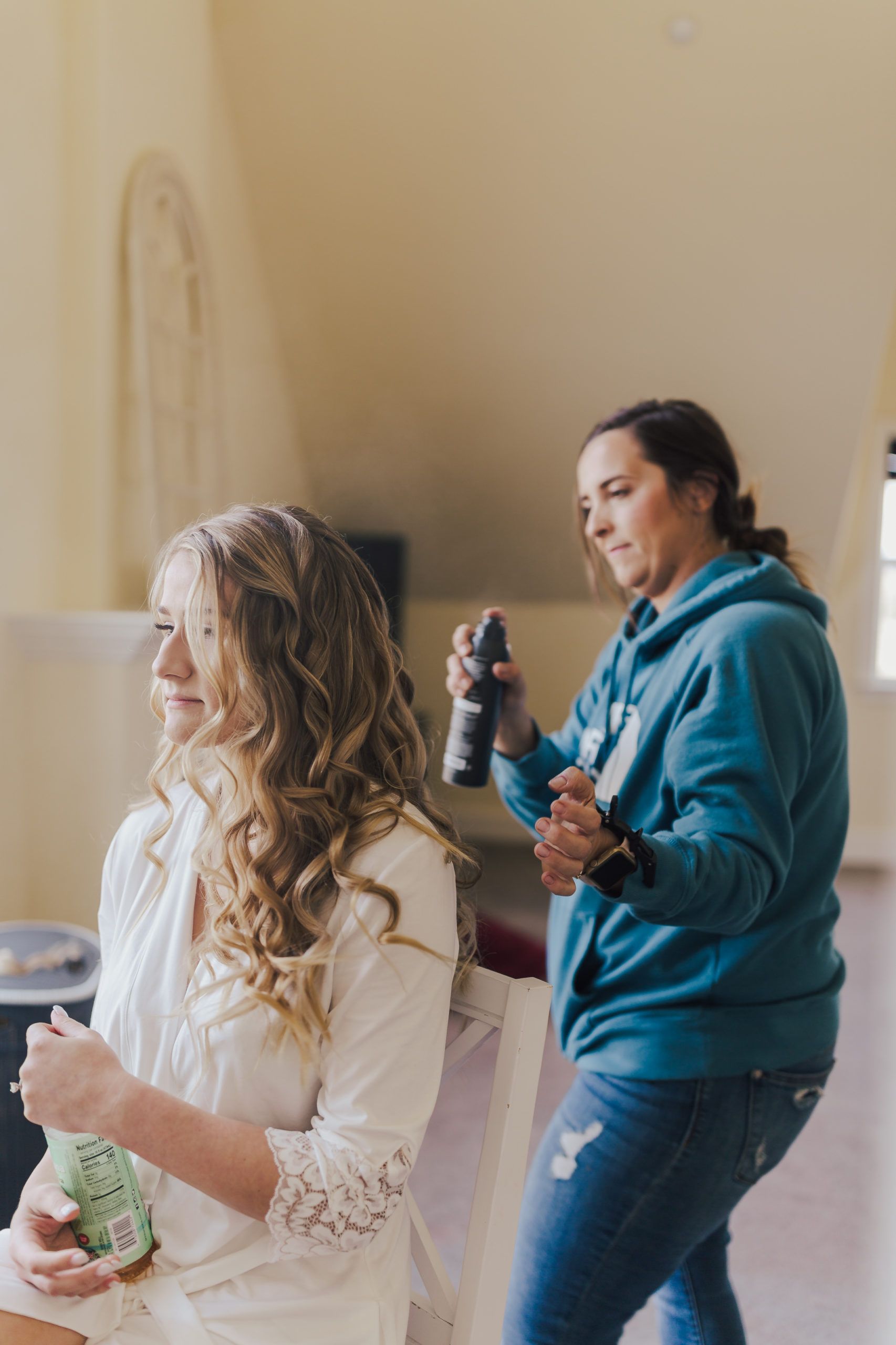 Woman has hair styled as another applies hairspray indoors.