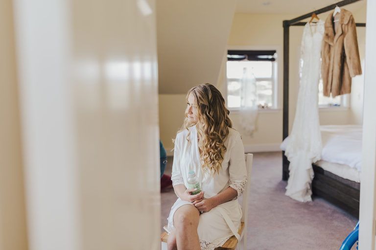 Woman in white robe looking away, wedding dress and coat hanging in a bedroom.