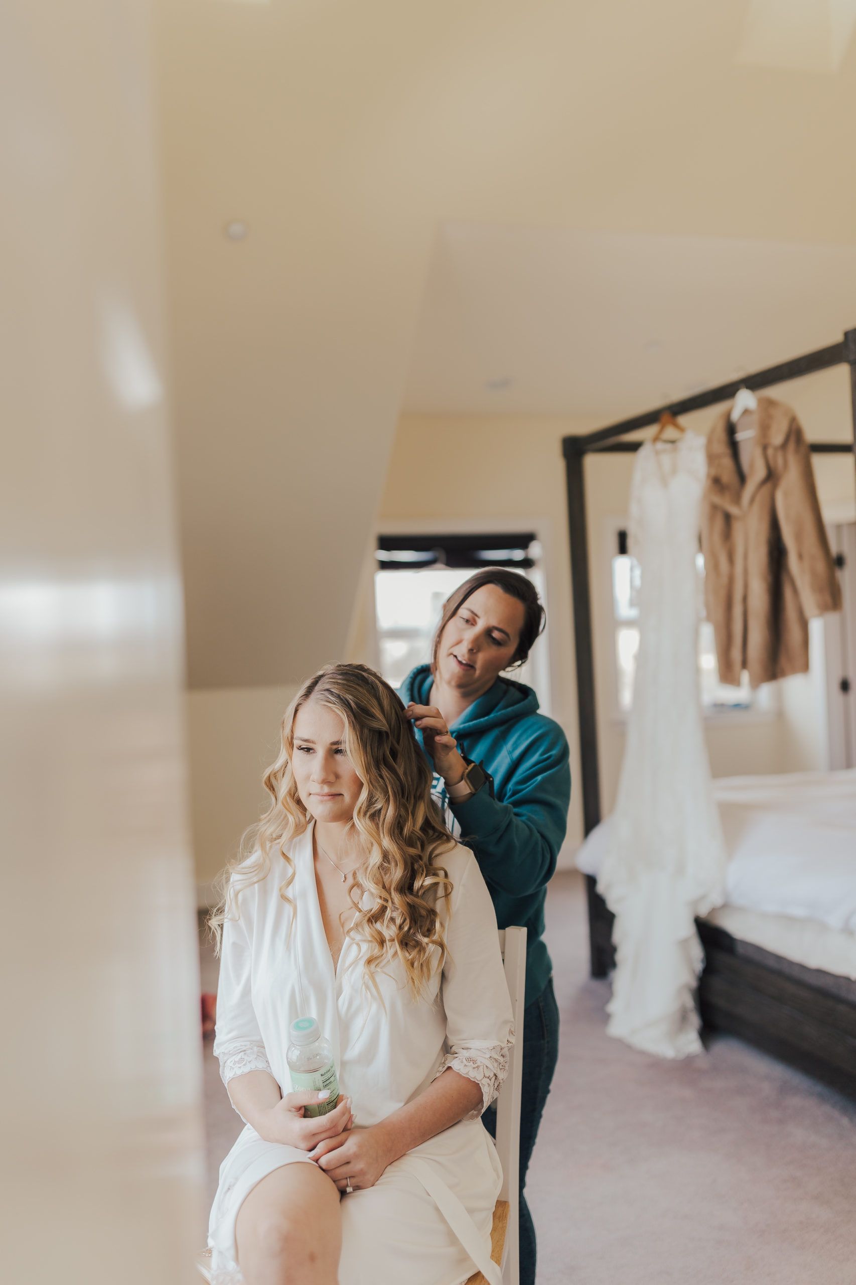Woman in a robe has hair styled by another woman in a teal sweatshirt; wedding dress and jacket hang in the background.