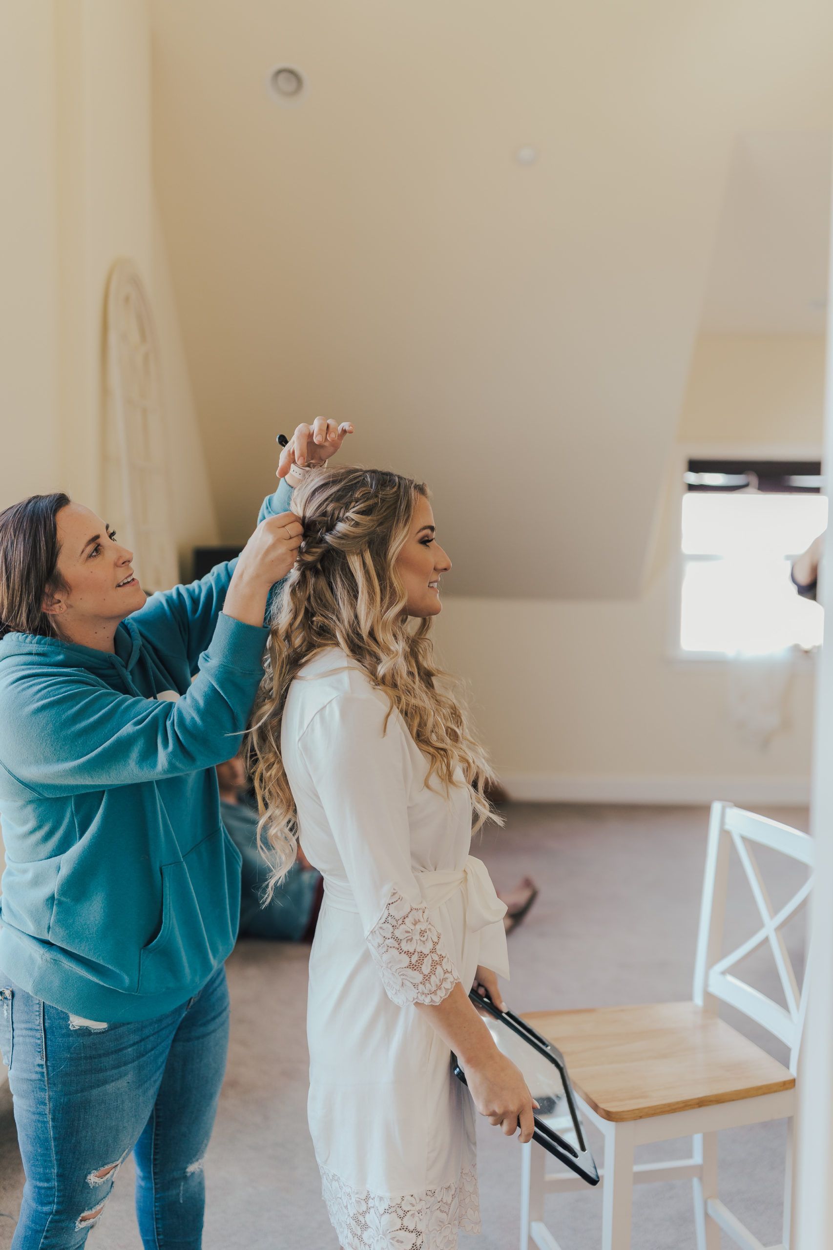 Woman having her hair styled by another woman in a white robe. Room has a window and a chair.