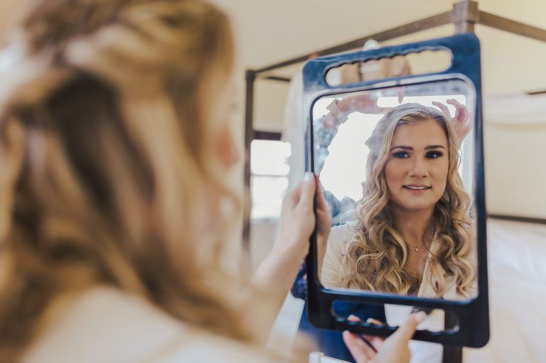 Woman looks in a mirror, adjusting her curly hair. Bedroom setting, neutral tones.