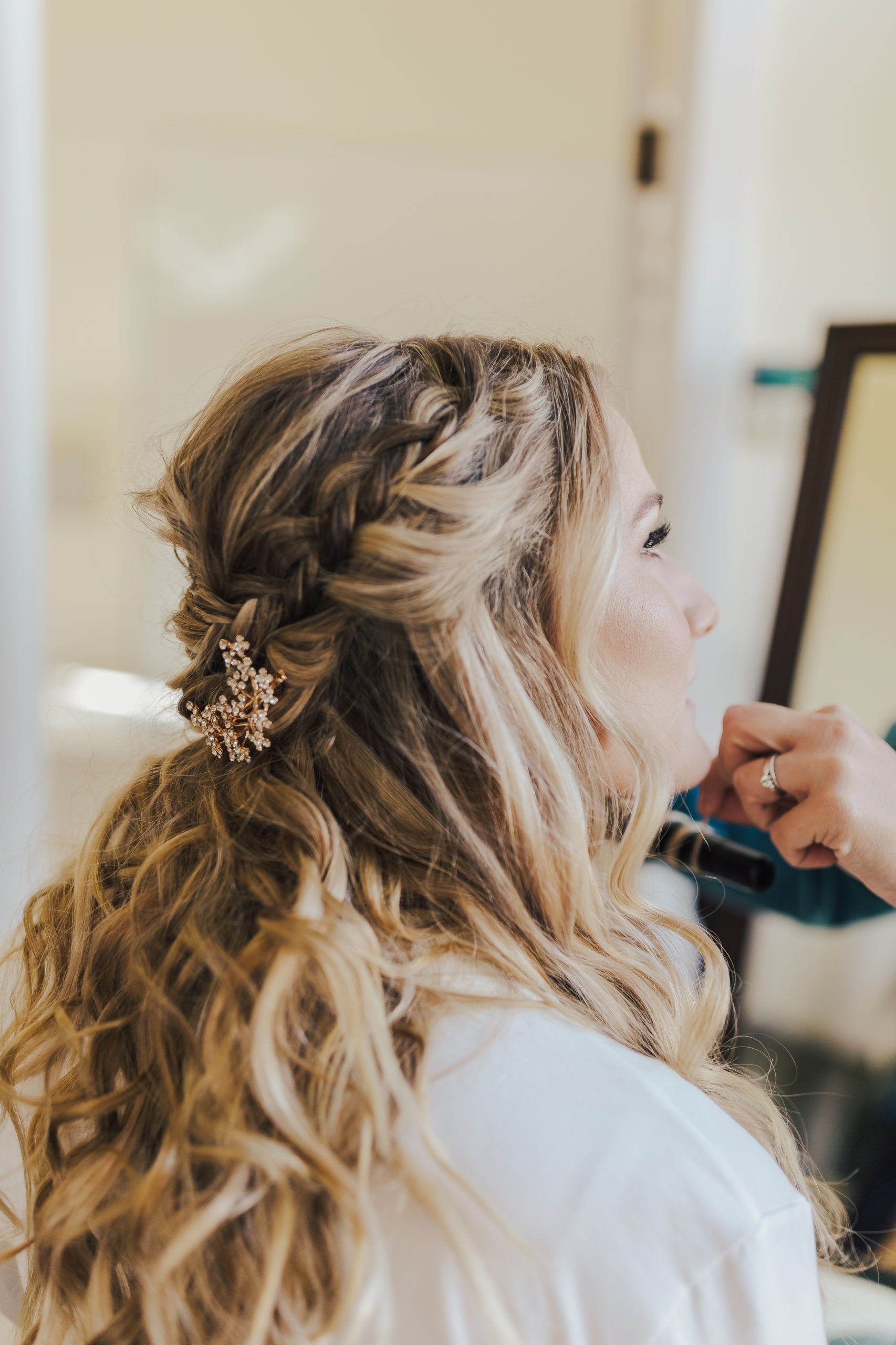 Woman with blonde hair in a braided updo, having makeup applied indoors, wearing a white robe.