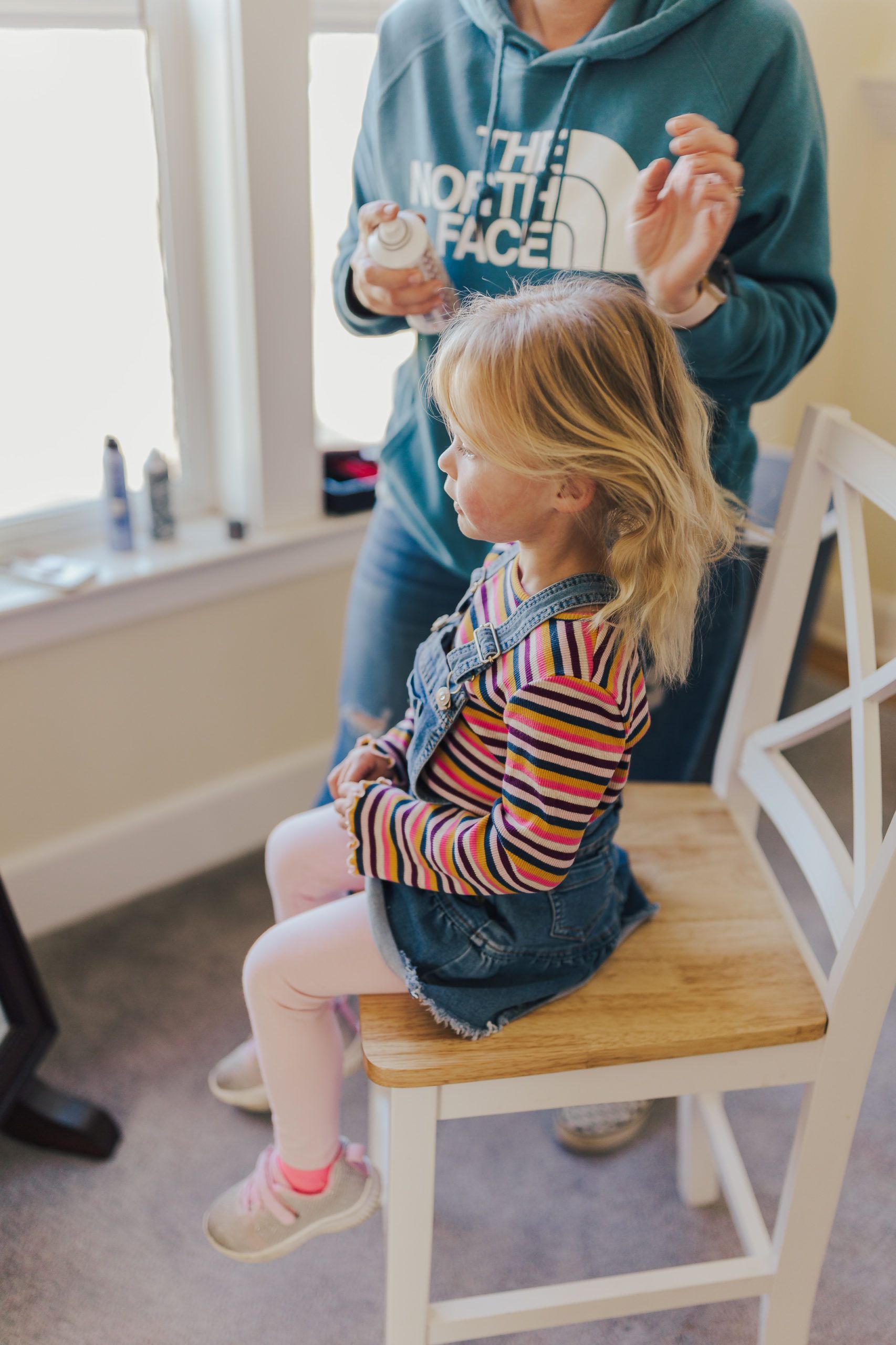 Girl seated on chair as an adult styles her blonde hair. Indoor setting, sunny window.