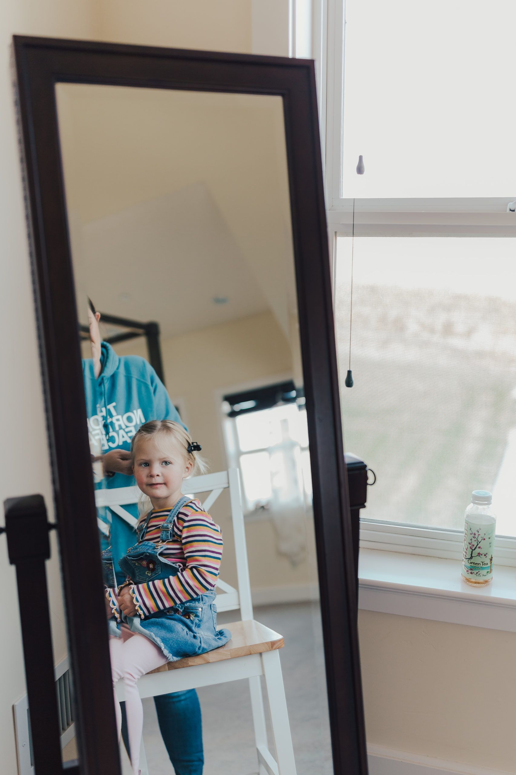 Girl with braided hair in overalls smiles, reflected in a full-length mirror.