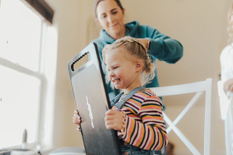 A young child smiles while holding a black board as an adult braids her hair indoors.