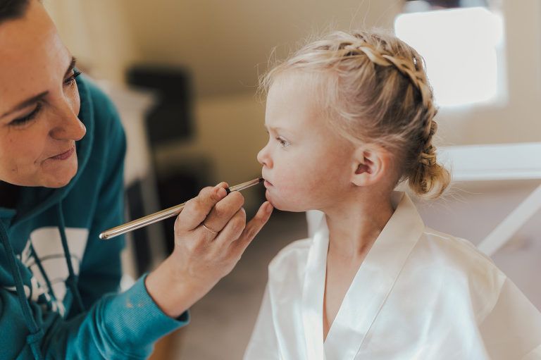 Woman applying makeup to a young child's lips; child in white robe, braided hair.