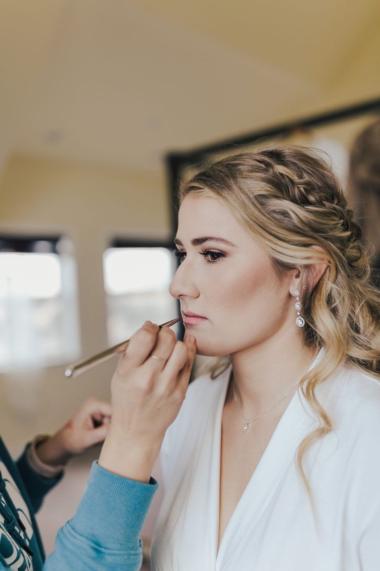 Makeup artist applying lipstick to a woman with blonde hair in a white robe, in a well-lit room.
