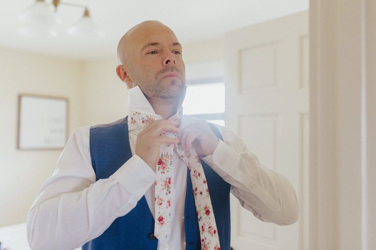 Man adjusting a floral tie in a room with white walls, wearing a blue vest and white shirt.