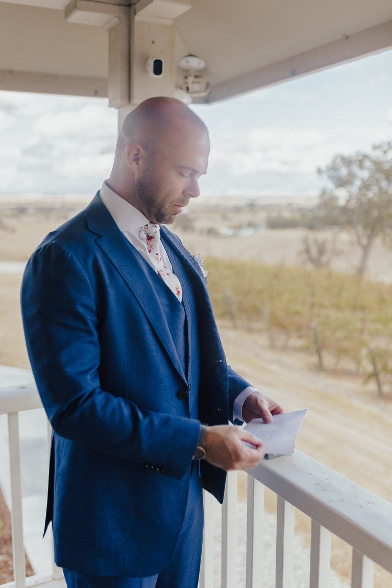 Man in blue suit, reading a letter on a balcony overlooking a rural landscape.