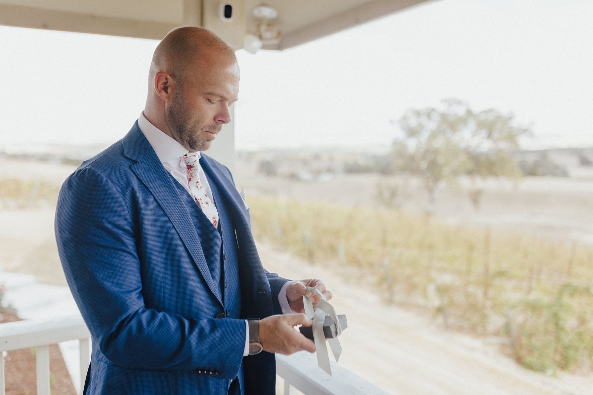 Man in blue suit, looking down, holding object on a balcony with a landscape view.