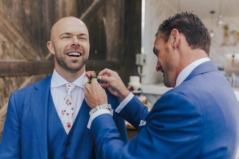 Man in blue suit smiling as another man adjusts his lapel flower. Rustic setting.