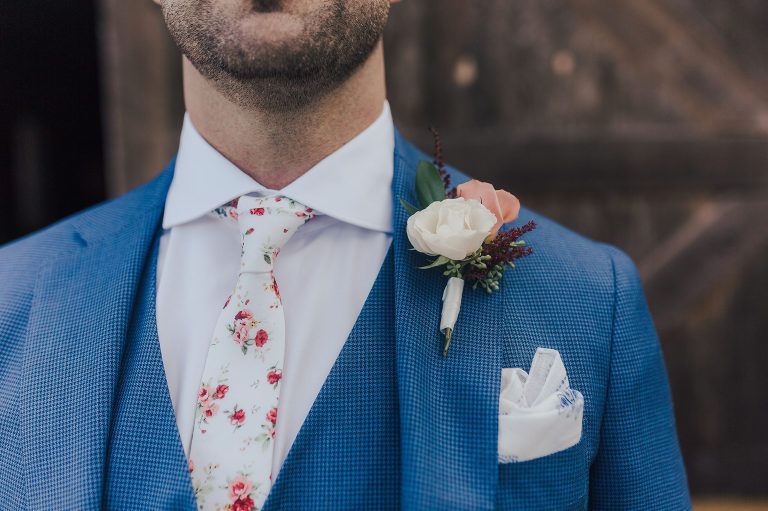 Man in blue suit, floral tie, and boutonniere; posing for a formal occasion.