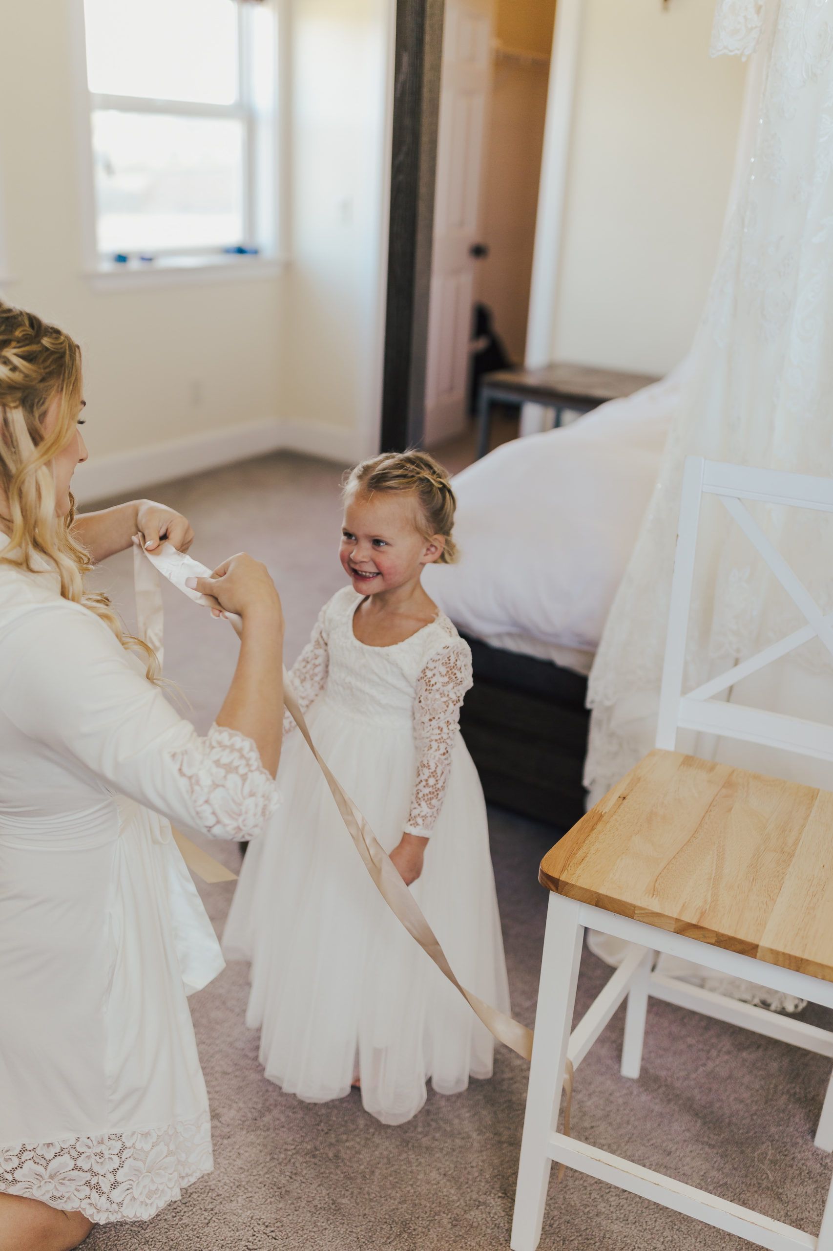 Woman in robe tying ribbon on flower girl's dress in a bedroom.