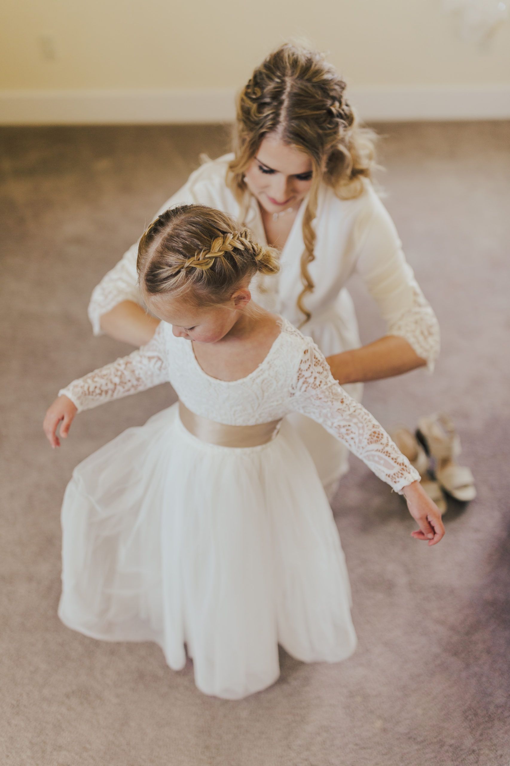 Woman adjusting a little girl's white dress, both with braided hairstyles.