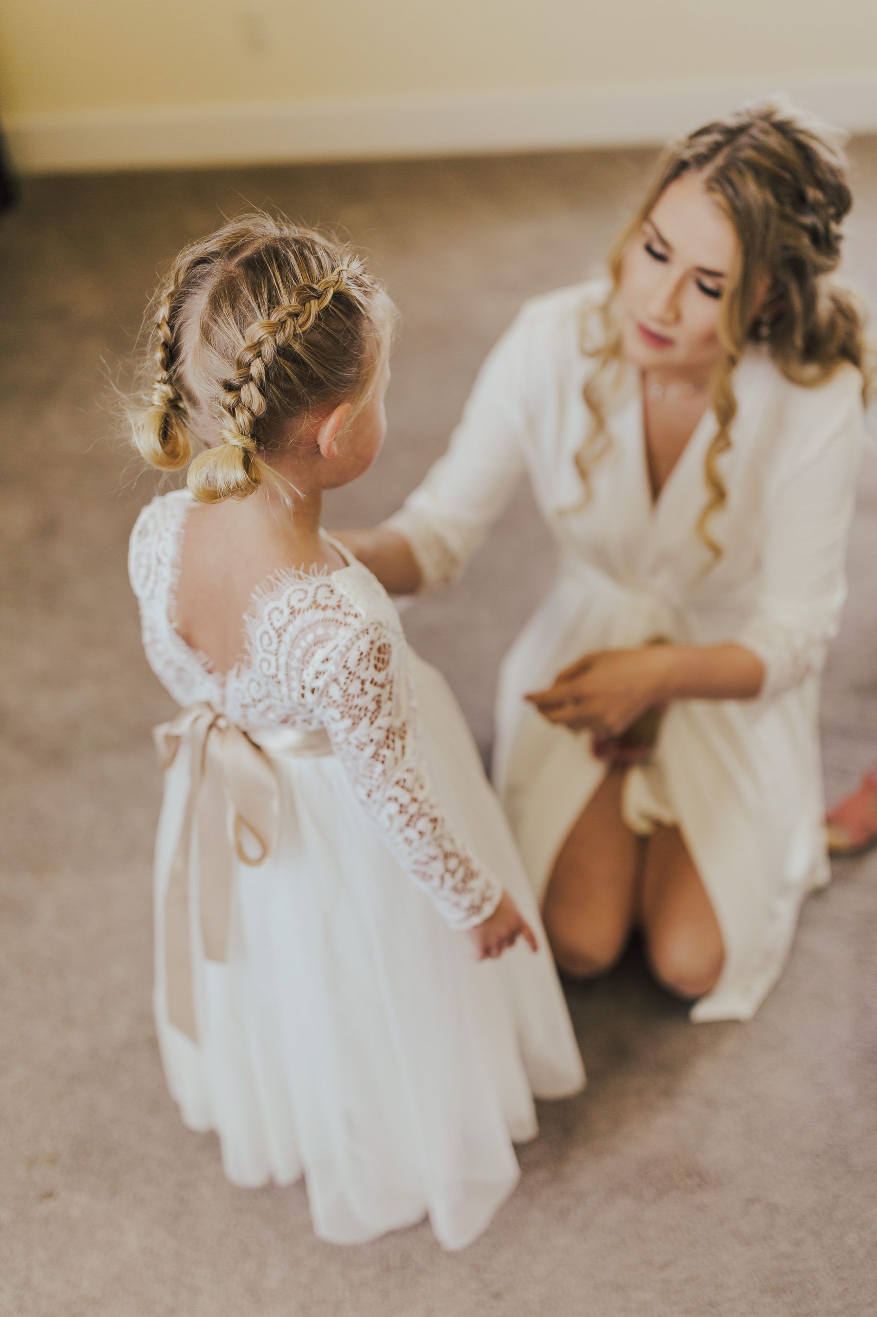 Flower girl in white dress with long sleeves, getting ready. Woman in robe kneels, adjusting dress.