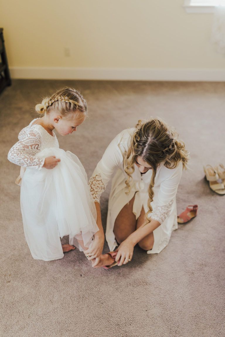 Woman helps flower girl with her shoe on a carpeted floor. Both are dressed in white.