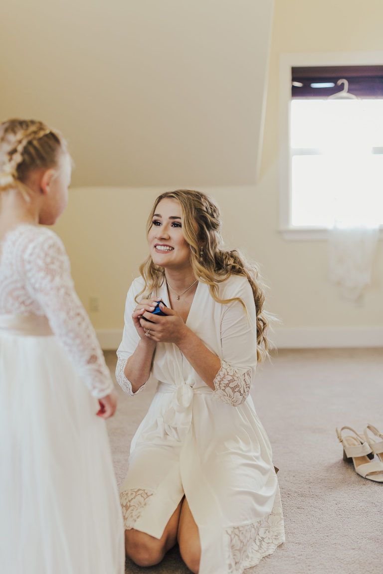 Woman kneeling in a robe, smiling at a flower girl, holding a small box, indoors.