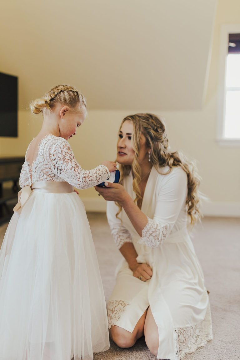 Woman kneeling, showing a ring box to a girl in a white dress with a braid. Room with a light color scheme.