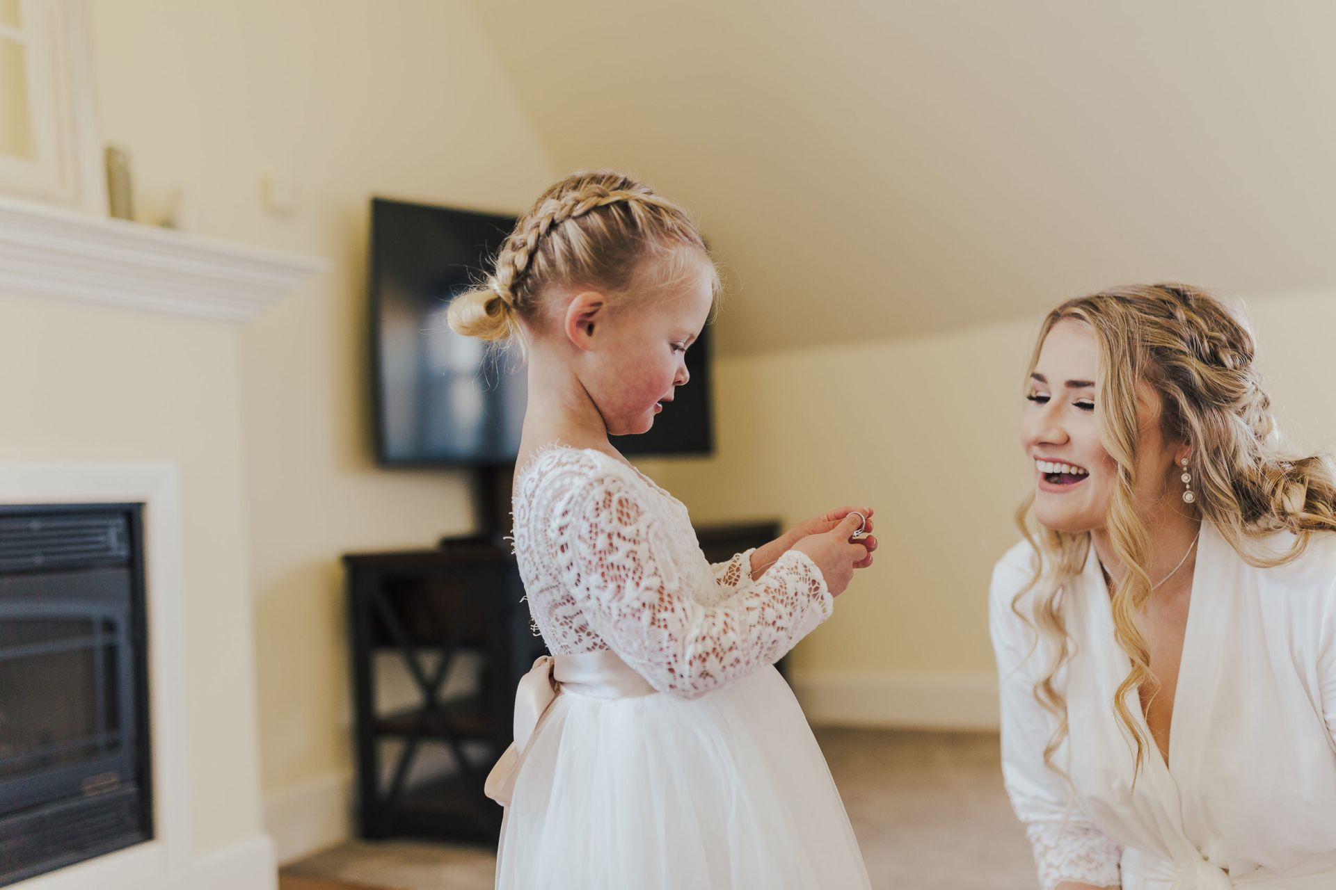 A young girl in a white dress and a woman in a white robe smile at each other indoors.