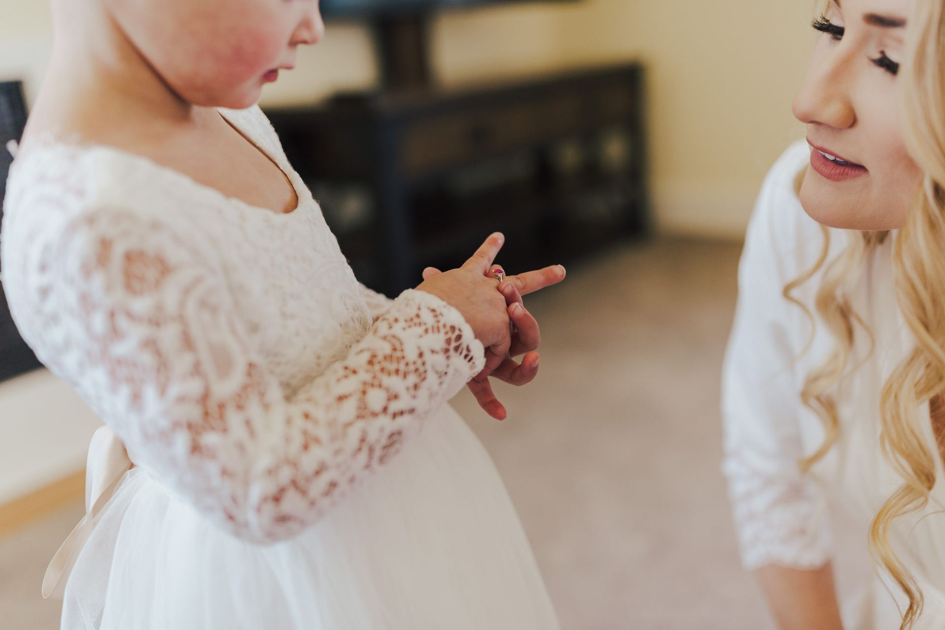 Girl in white dress examines her hands as woman looks on. Indoor setting.