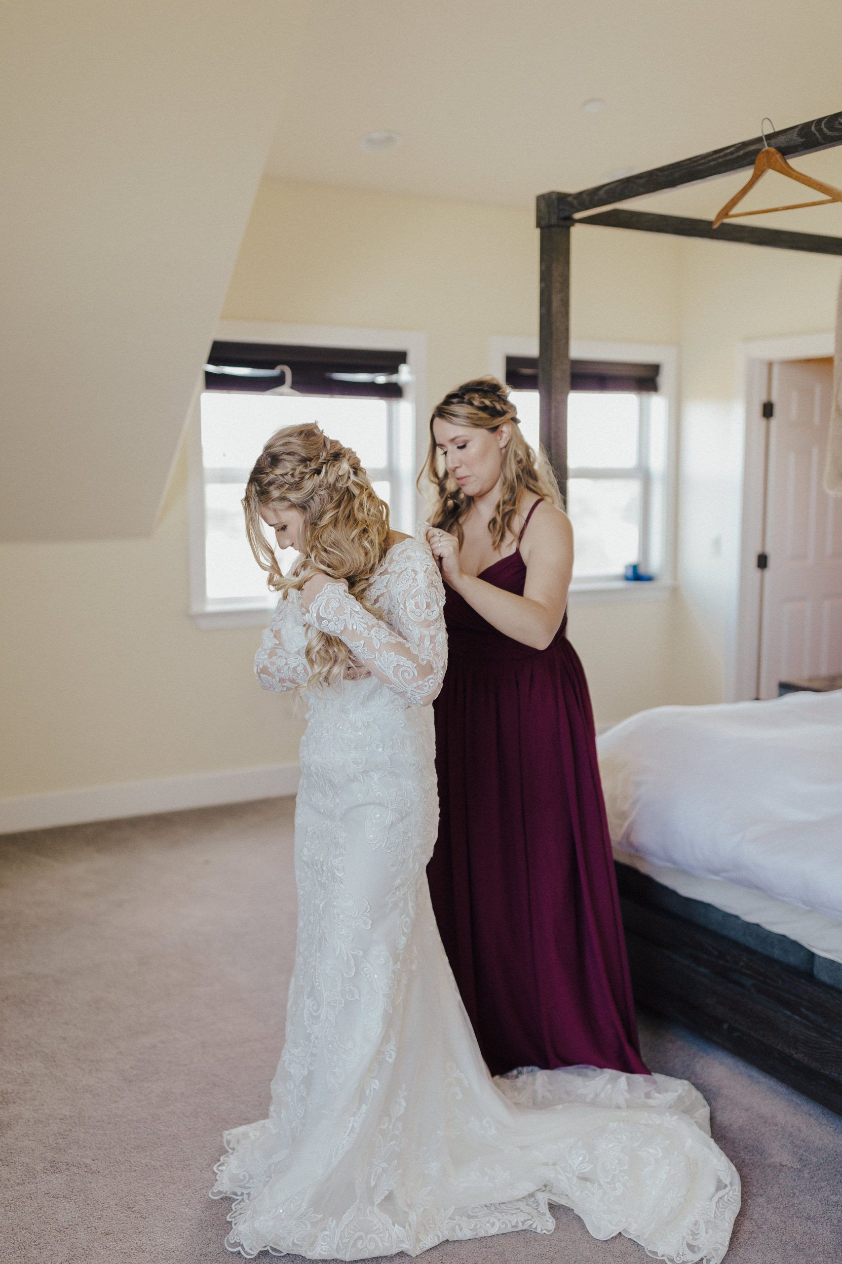 Bride adjusting wedding dress as bridesmaid assists in a bedroom.