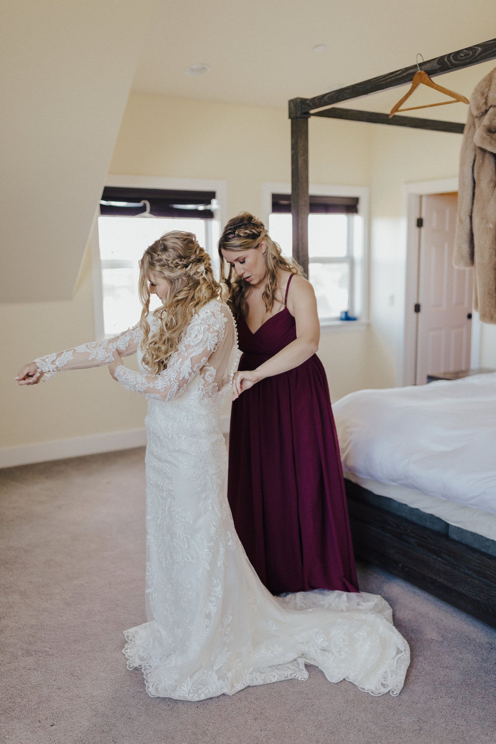 Bride getting help with dress by bridesmaid in a bedroom. The dress is white lace.