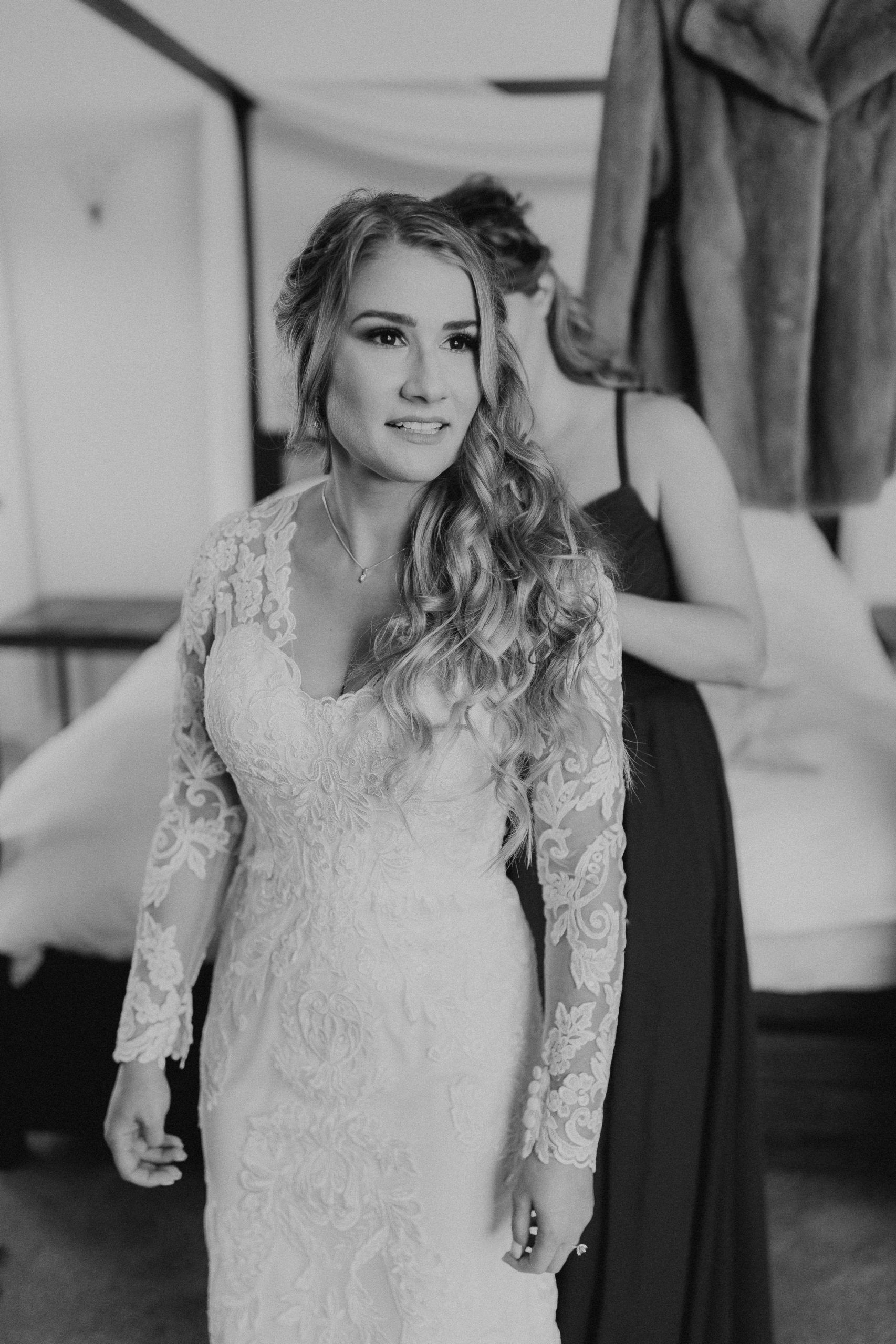Bride in lace dress, hair styled, being helped by bridesmaid in a room with soft lighting.
