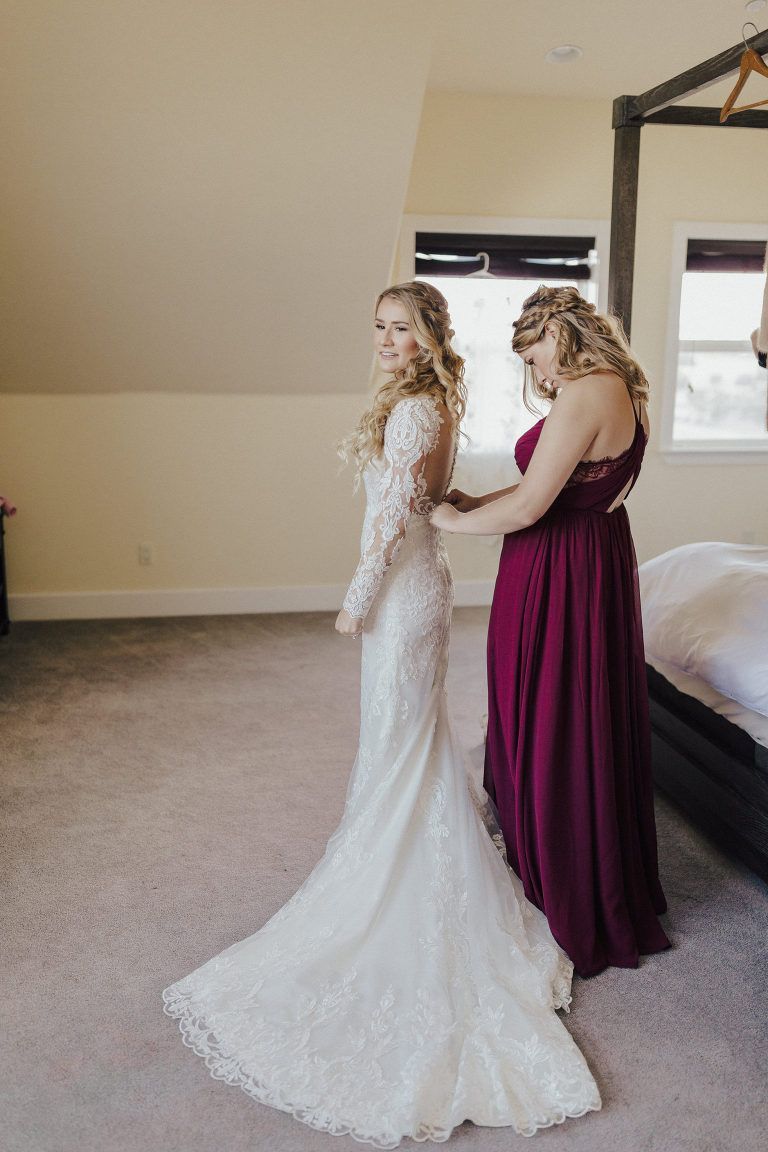 Bride in lace gown, being helped by bridesmaid in burgundy dress in a bedroom.
