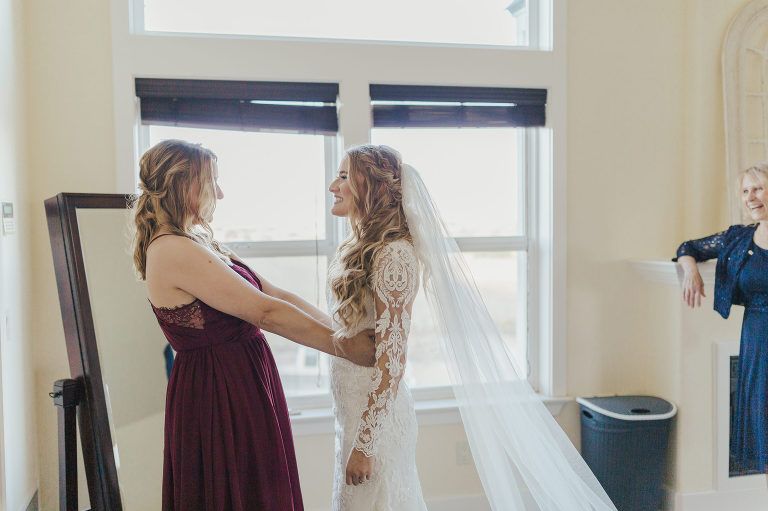 Woman in wedding dress being helped by another, near window. Third woman watches.