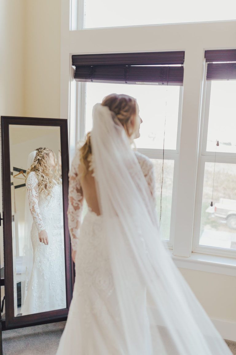 Bride in white lace dress and veil, facing window, looking in mirror.